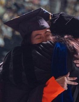 Graduate in cap and gown at ceremony in heartfelt embrace with faculty member